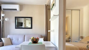 A compact living space with a sofa, a small dining table with a bowl of fruit, a white chair, an air conditioning unit, and a bed with a polka dot blanket.