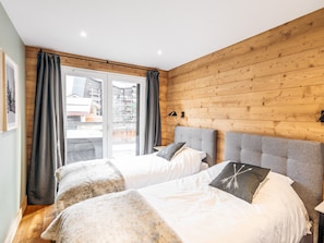 A bedroom with two beds, wooden walls, a large window with curtains, and a view of buildings outside.
