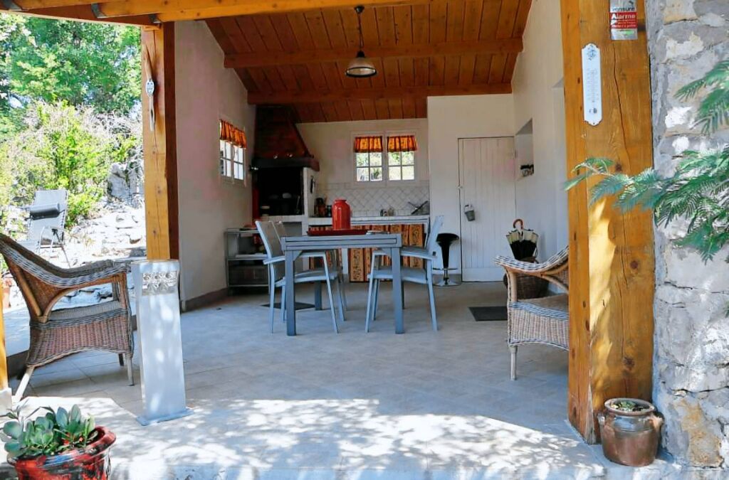 Un'area pranzo esterna riparata con tavolo e sedie, un muro in pietra e vista sulla cucina sullo sfondo.