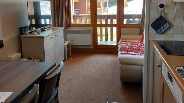 Una habitación compacta con escritorio de madera, una silla, cama con manta roja y blanca, pequeña kitchenette y puerta corrediza de vidrio que da a un balcón exterior.