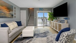 A living room with a sofa, ottoman, and a flat-screen TV. There is a staircase leading to a balcony with outdoor furniture.