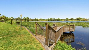 A wooden boardwalk over water with a grassy area and lamp posts on the left.