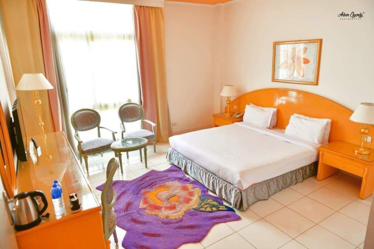 Una habitación de hotel con una cama, dos sillas, una mesita con una tetera y una ventana con cortinas.