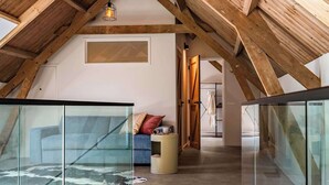 Ett rum i loftstil med synliga träbjälkar, ett glasbord med en matta av kohud, en blå soffa och ett litet runt bord med en kopp på.