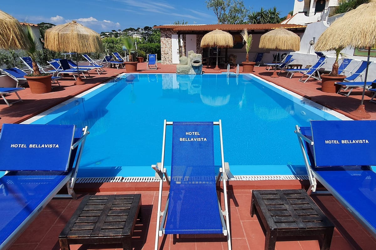 Une piscine entourée de chaises longues bleues et de parasols, le tout bordé par un bâtiment doté d’un toit en tuiles et de parasols en chaume.