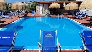 A hotel pool with clear blue water, surrounded by blue lounge chairs and umbrellas, and a stone structure in the background. - Hotel Bellavista (Forio)