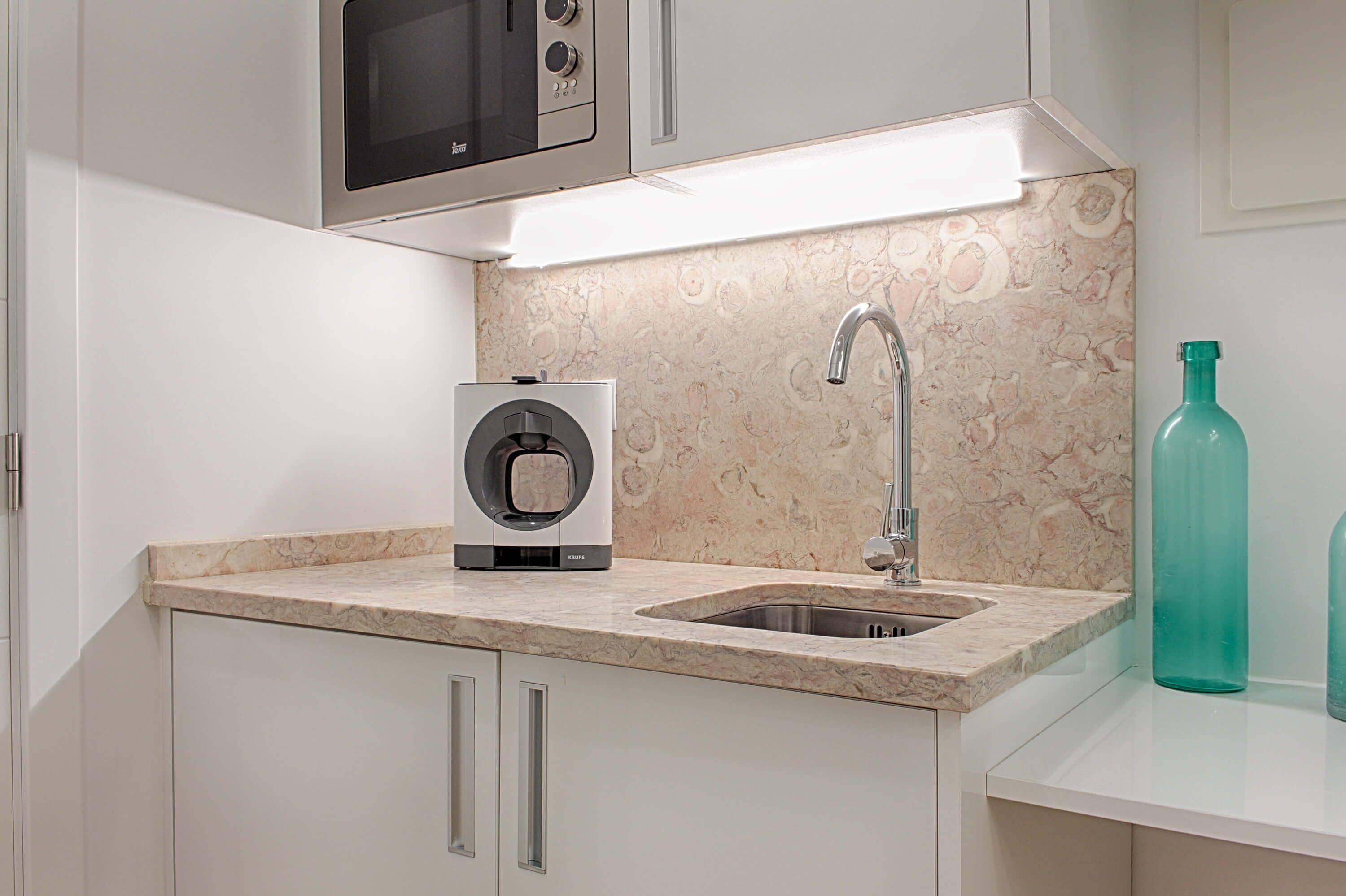 A kitchen with a built-in microwave, a coffee machine, a sink, and a marble backsplash.