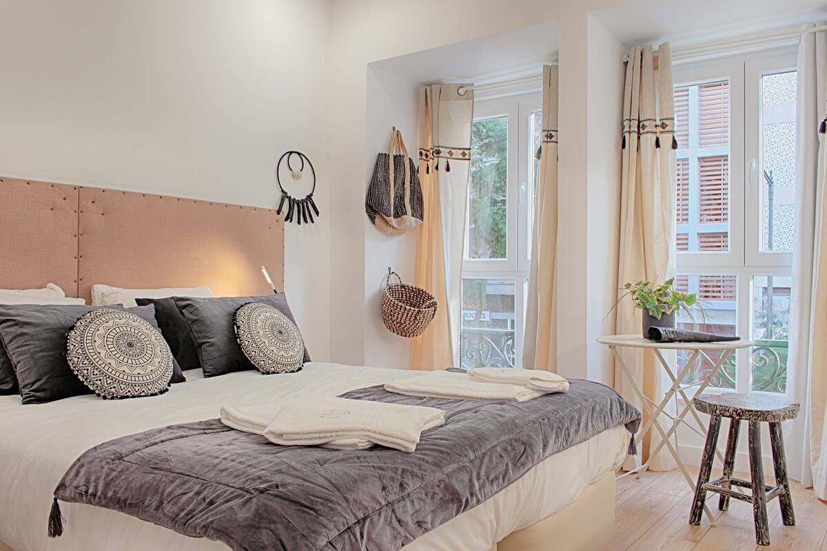A bedroom with a large bed, a bedside table with a plant, and a window with curtains.