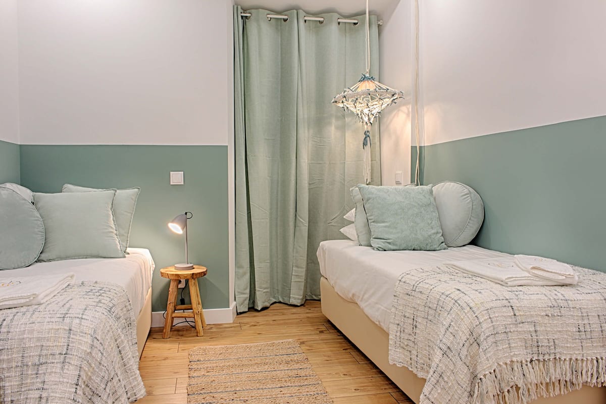 A bedroom with two beds, a small stool, a hanging light fixture, and a curtain.