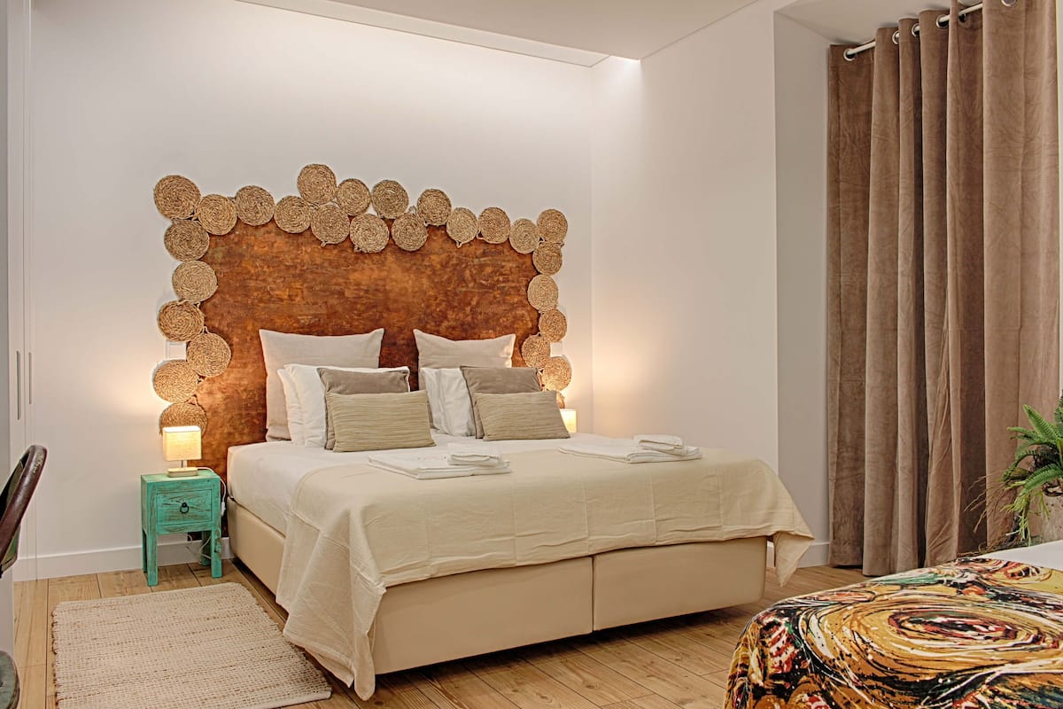 A bedroom with a unique woven headboard, a bed with beige bedding, a small teal nightstand with a lamp, and a large window with curtains.