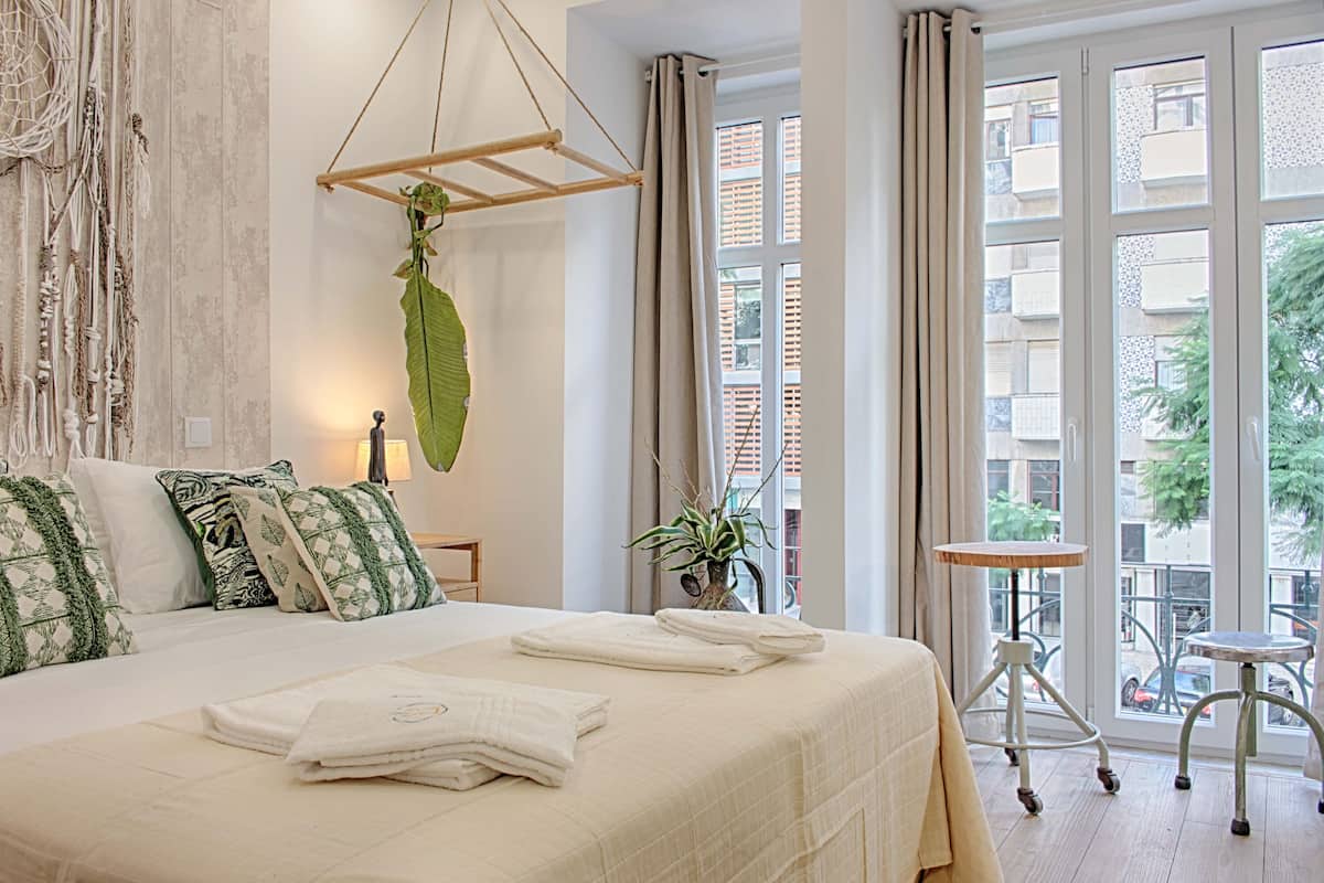 A neatly made bed with white linens and green patterned pillows, a hanging plant, and a view of the city through large windows.
