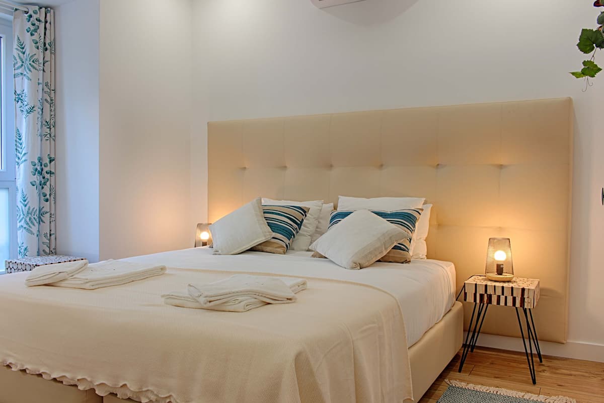 A neatly made bed with white linens and striped pillows, a bedside lamp, and a window with curtains.