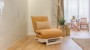 A room with a wooden floor, a white wardrobe, a beige armchair, a small table with chairs, and a large window with sheer curtains. - GuestHouse by Trip2Portugal (Lisbon)