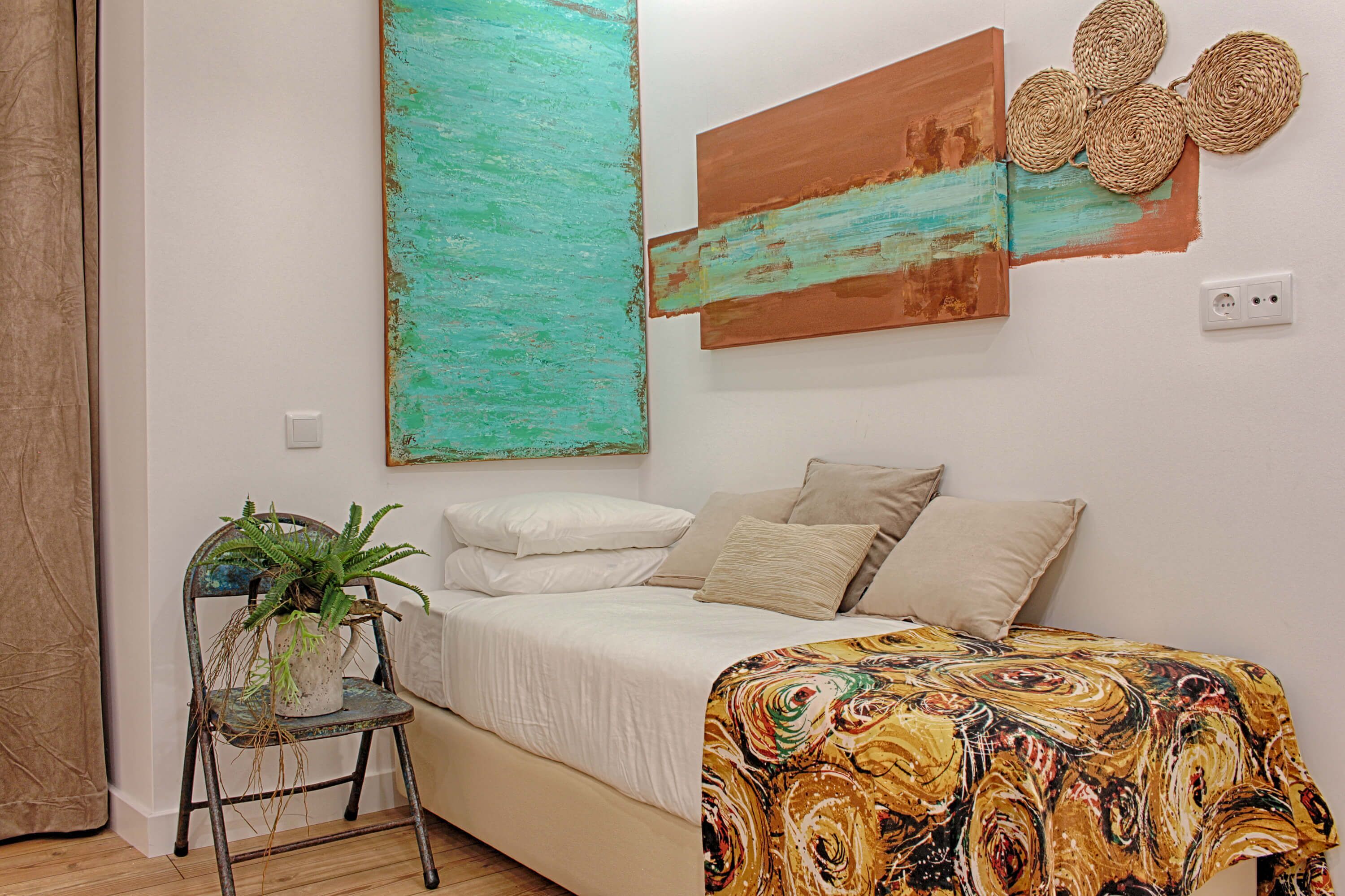 Une chambre à coucher avec un lit, une chaise agrémentée d’une plante et deux tableaux abstraits accrochés au mur.