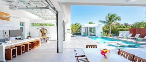 A modern kitchen with a long counter and stools, a dining area with a table and chairs, and a view of a swimming pool and palm trees.