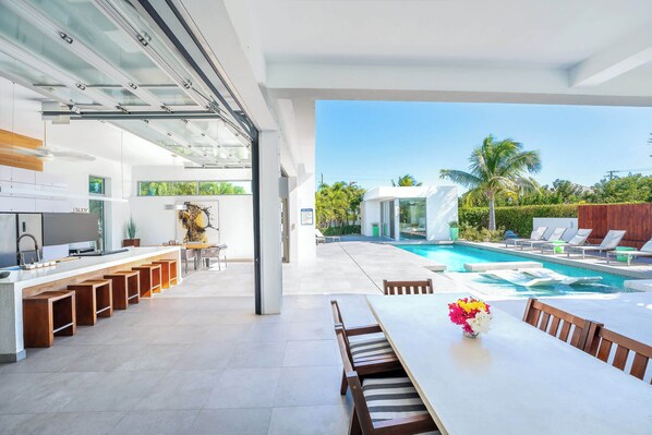 A modern kitchen with a long counter and stools, a dining area with a table and chairs, and a view of a swimming pool and palm trees.