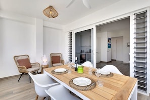 A dining area with a wooden table set for four, surrounded by wicker chairs, a hanging light fixture, and a view into the kitchen.