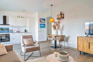 Un salon moderne avec un canapé, un coin repas comprenant une table et des chaises, une cuisine avec des meubles blancs et un téléviseur posé sur un meuble en bois.