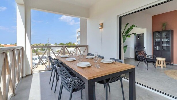 A dining area with a wooden table set for four, surrounded by chairs, with a view of the sky and buildings outside.