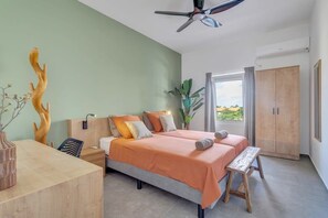 Ett sovrum med en säng, sängbord, en träpanel som huvudgavel, en takfläkt, ett fönster med gardiner och utsikt över grönområden utanför.
