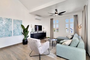 A modern living room with a sofa, a chair, a small table, and a TV. There are two large windows with curtains, a ceiling fan, and artwork on the walls. - Kavel 17 - Laguna Breeze with a shared pool and spectacular views of the Lagune (Kralendijk)