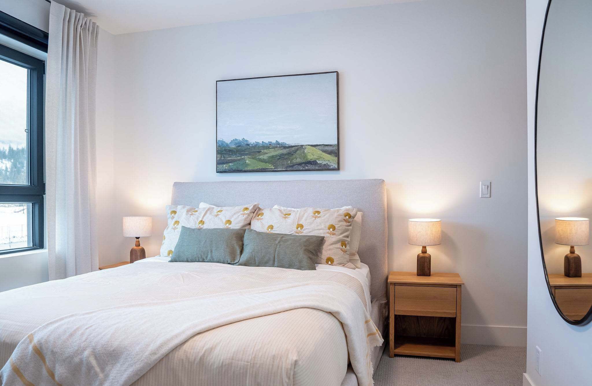 A bedroom with a bed, two bedside tables with lamps, a wall-mounted picture, and a window with curtains.