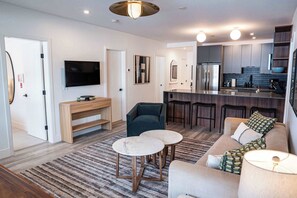 Uma sala de estar moderna com um sofá, duas mesas redondas, uma TV e uma cozinha com geladeira e banquetas.