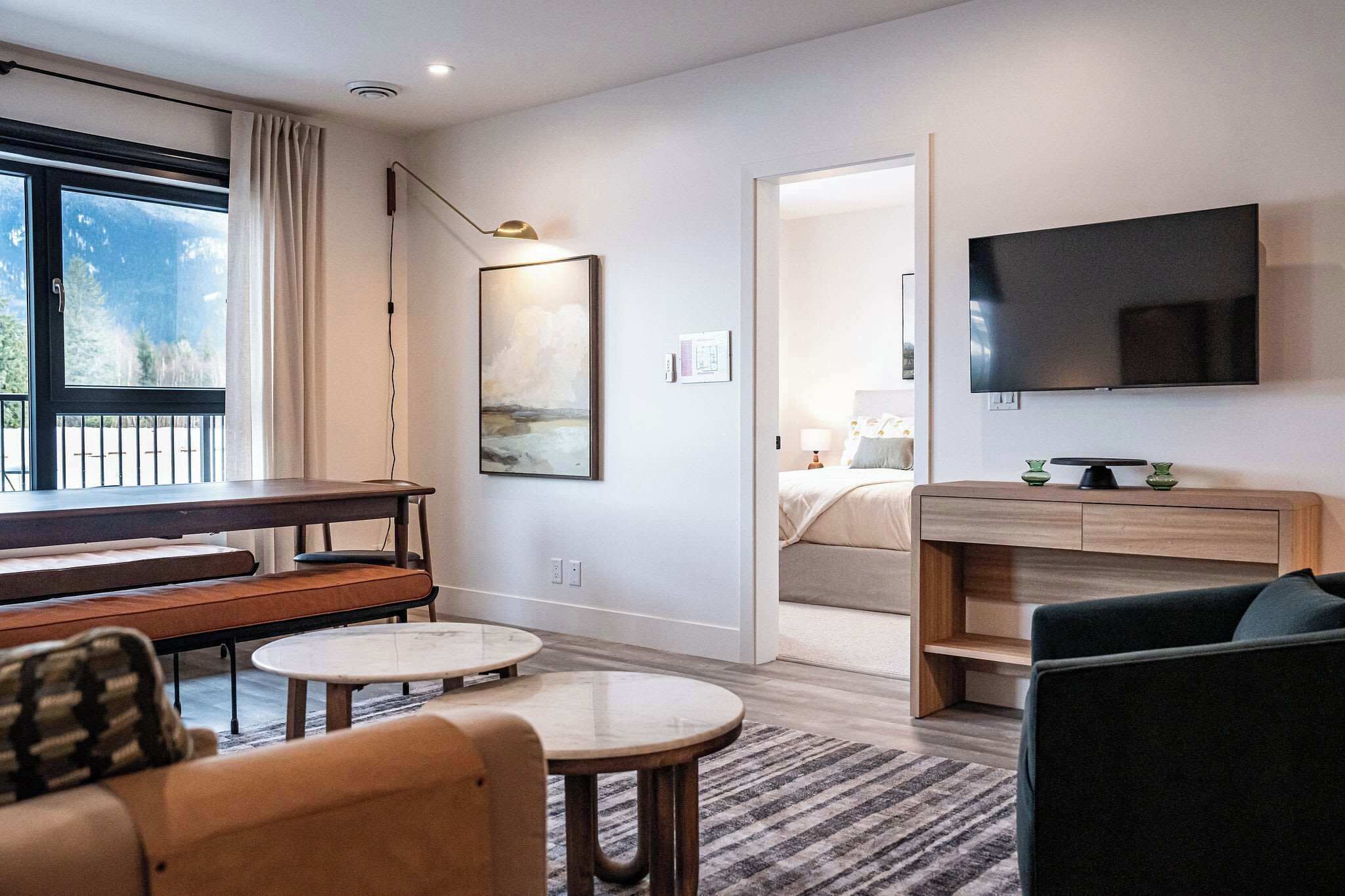 Una habitación de hotel moderna con un ventanal grande, un televisor de pantalla plana, una zona de comedor con un banco y una cama con una mesita de noche.