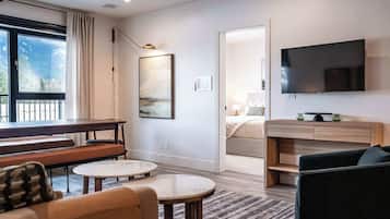 Una habitación de hotel moderna con un ventanal grande, un televisor de pantalla plana, una zona de comedor con un banco y una cama con una mesita de noche.
