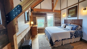 Ein Schlafzimmer mit Holzbalkendecke, einem Bett mit gemusterten Betttextilien, einem Nachttisch, einer Lampe und einem Fenster mit Blick ins Grüne.
