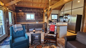 Ein gemütliches Blockhaus-Interieur mit einem Wohnbereich, der einen blauen Sessel, einen Kamin und einen Esstisch umfasst. Im Hintergrund ist eine Küche mit modernen Geräten und einem großen Fenster erkennbar.