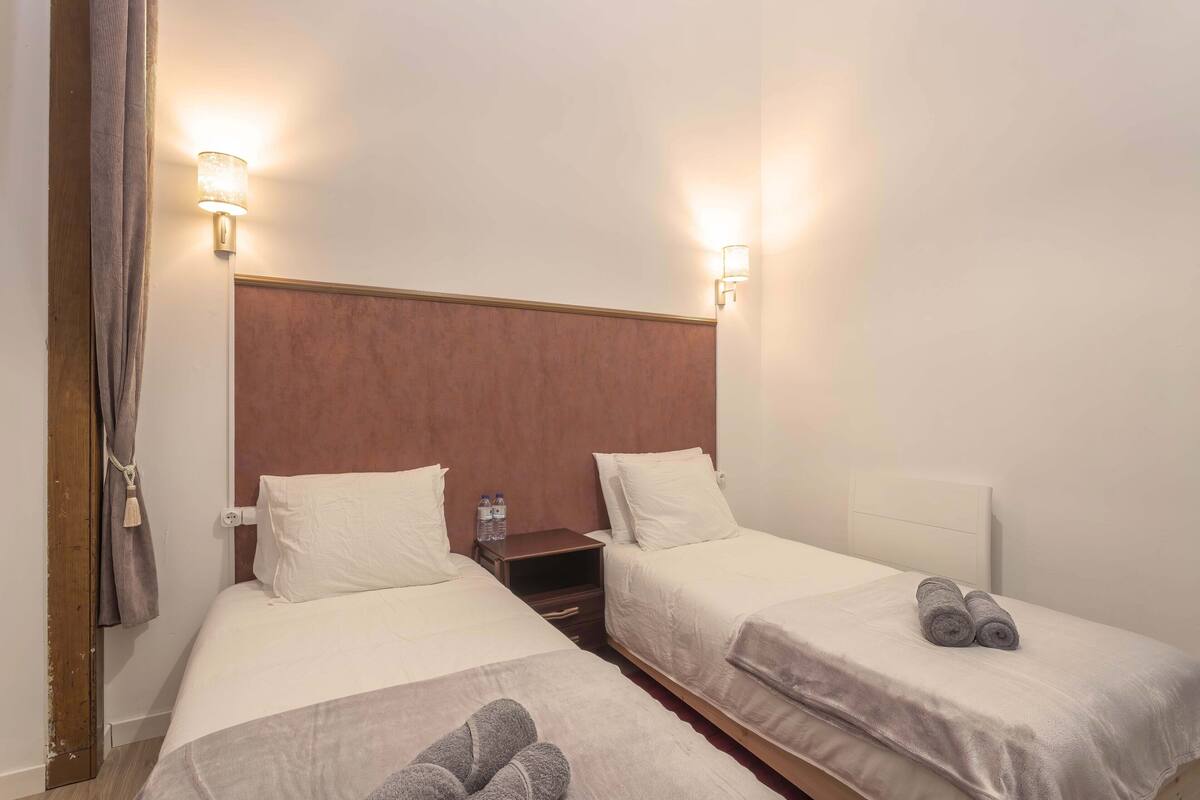Quarto de hotel com duas camas, cada uma com lençóis e almofadas brancos, cabeceiras de madeira e mesas de cabeceira com garrafas de água.