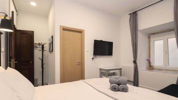 Quarto de hotel com cama, porta de madeira, televisão na parede, janela com cortinas e uma pequena mesa com uma planta.