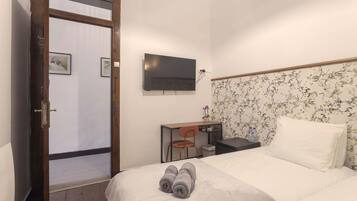 Quarto de hotel com uma cama, uma escrivaninha com cadeira, uma televisão na parede e uma porta que dá para outro cômodo.