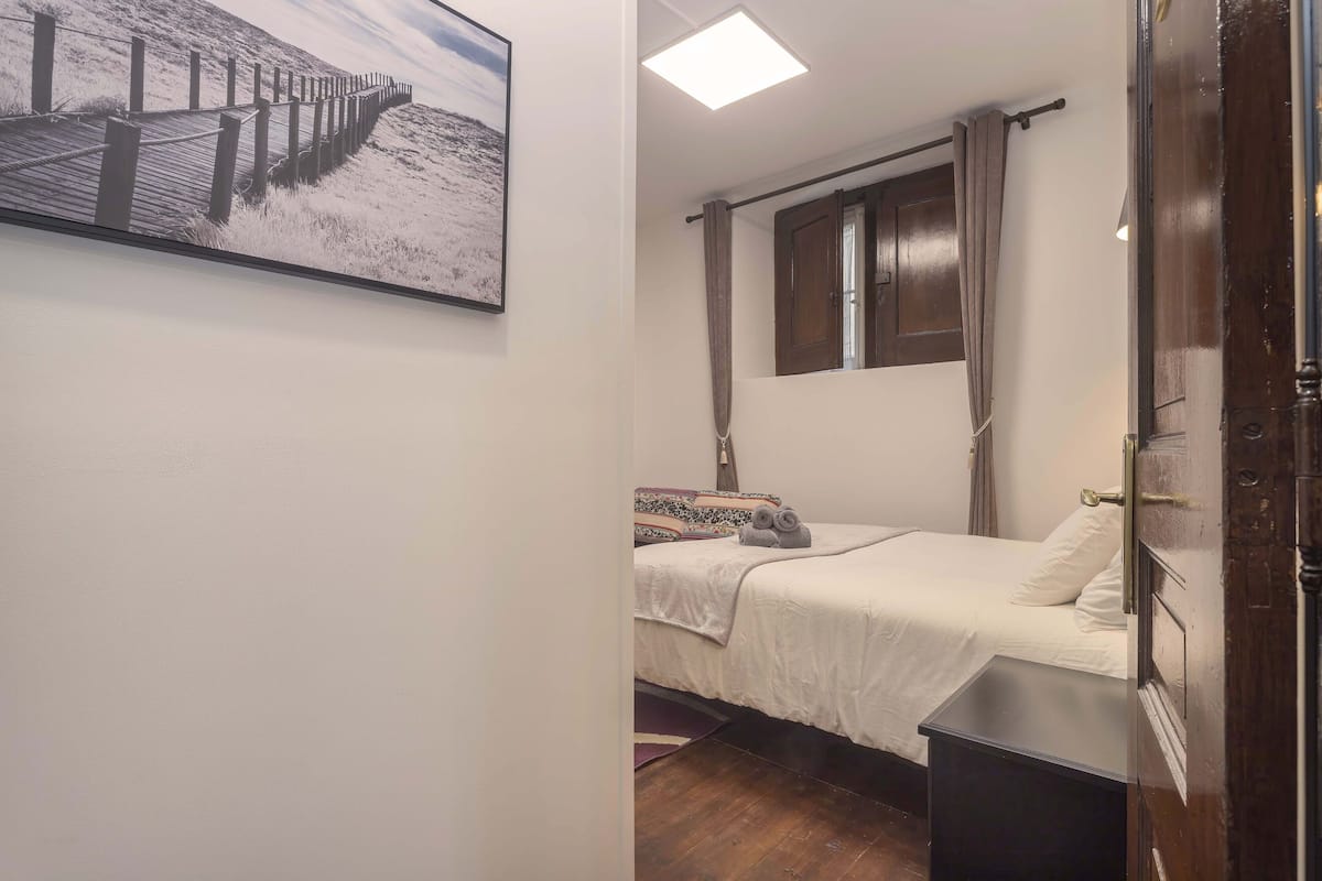 Quarto com cama, janela com cortinas, porta de madeira e um quadro na parede.