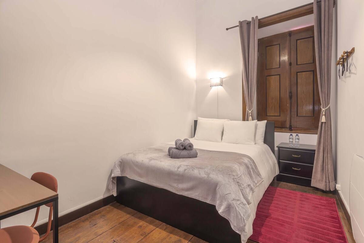 Quarto com cama, mesa de cabeceira, luminária, cadeira e tapete vermelho.