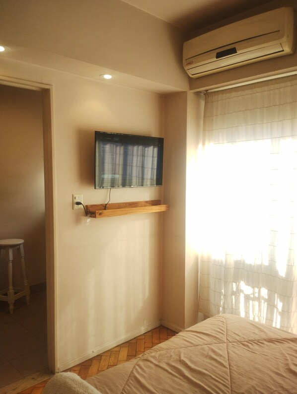 A bedroom with a bed, a mounted TV, an air conditioning unit, and a window with sheer curtains.