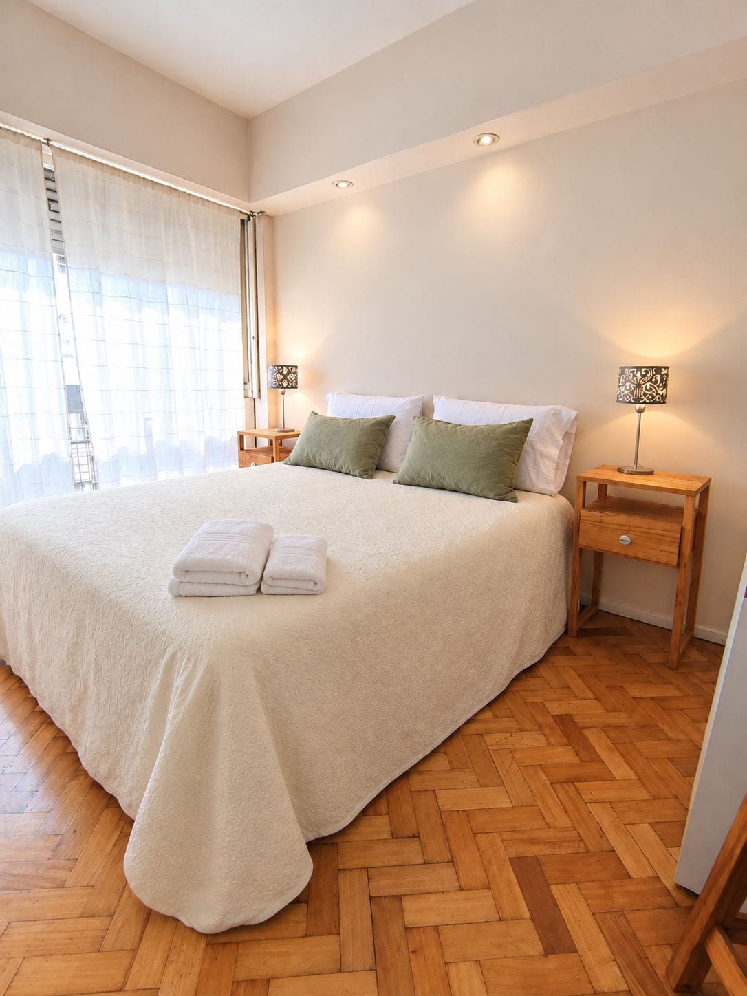 A neatly made bed with a white duvet and two pillows, a wooden nightstand with a lamp, and a window with sheer curtains.