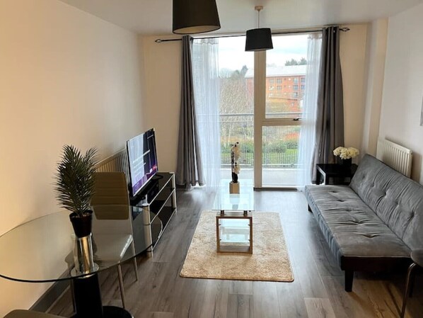 A modern living room with a sofa, TV, glass table, and a view of a building through large windows.
