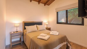 Ein Schlafzimmer mit Holzboden, einem großen Bett mit zwei Kissen, Nachttischen mit Lampen und einem Fenster mit Blick ins Grüne.