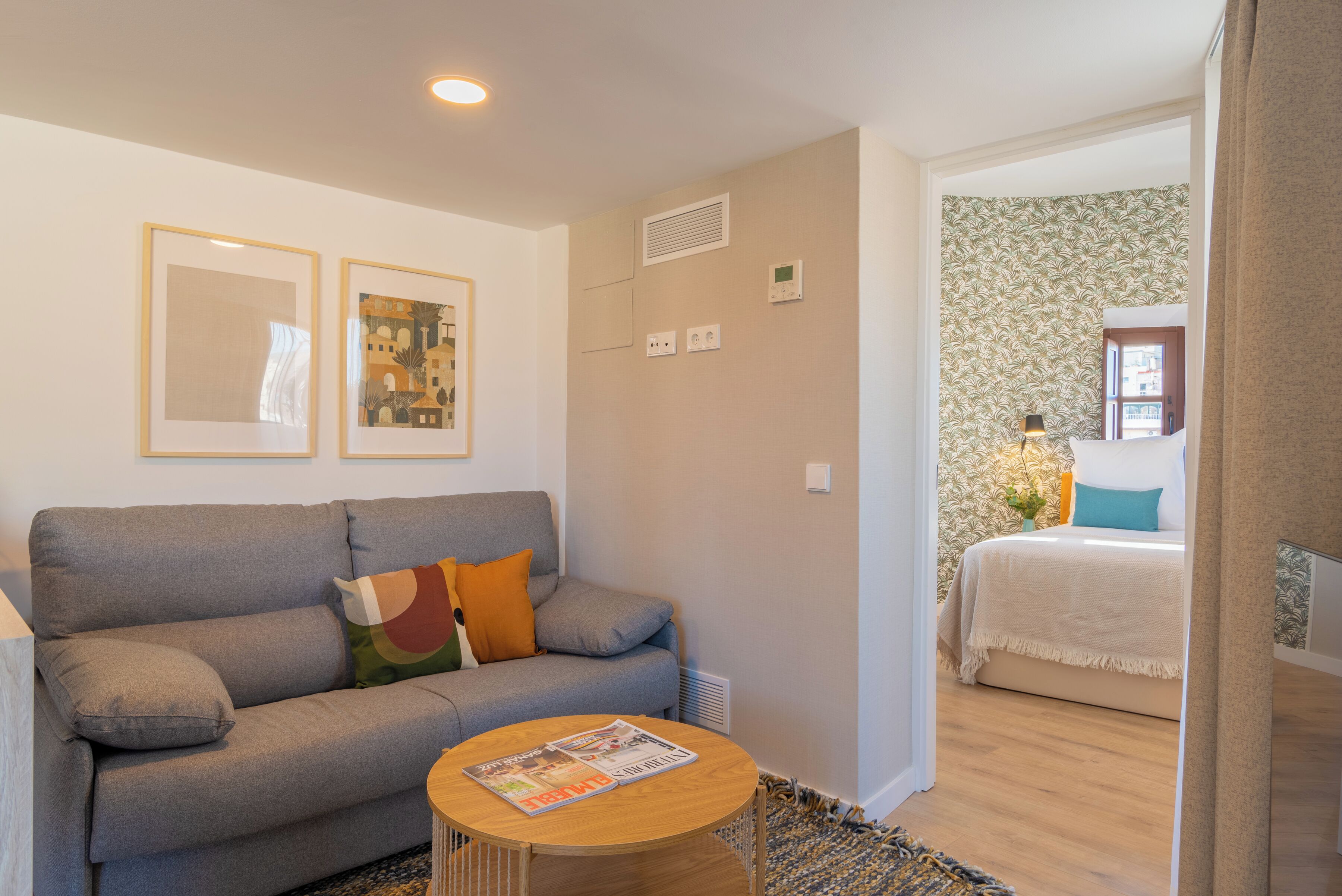 A living room with a grey sofa, a round wooden coffee table with magazines, and a patterned rug. There is a bed with a white duvet and teal pillows in the adjacent room.