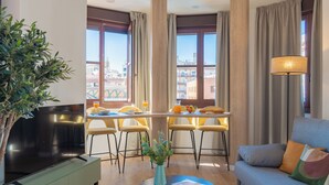 A modern living room with a flat-screen TV, a sofa, a dining area with yellow chairs, and a view of buildings through large windows. - Feelathome Atarazanas Apartments (Málaga)