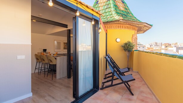 A balcony with a yellow wall, a green-roofed structure, and a sliding glass door leading to a dining area with chairs and a table. - Feelathome Atarazanas Apartments (Málaga)
