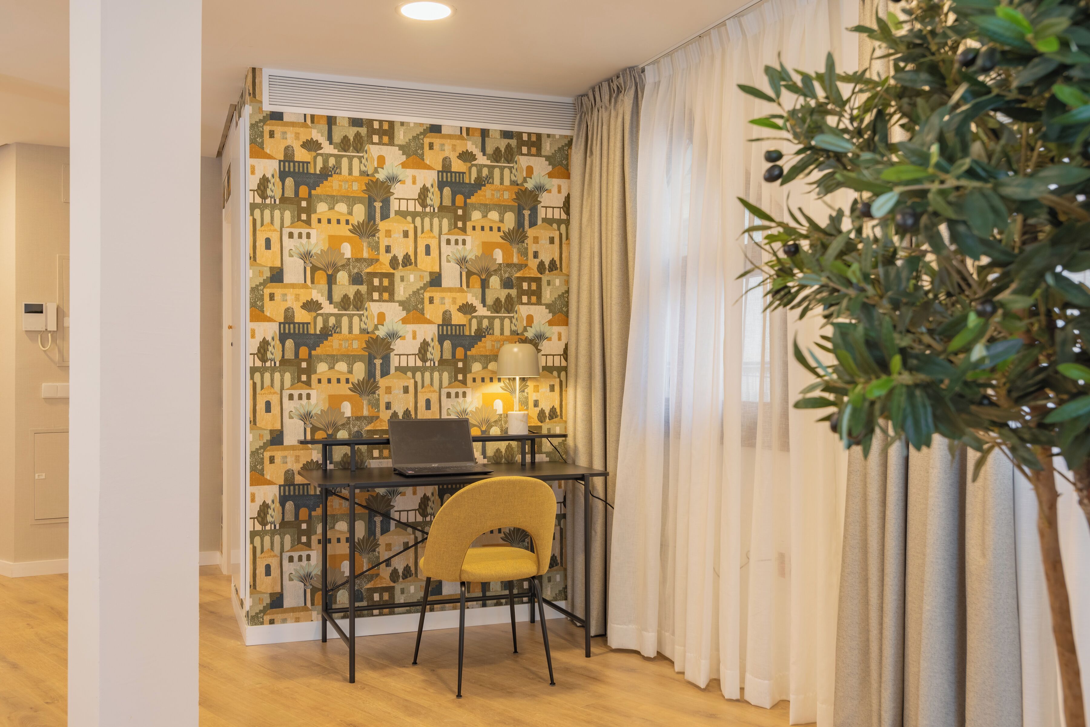 Een kamer met behang met een patroon, een bureau met een laptop, een gele stoel en een plant.