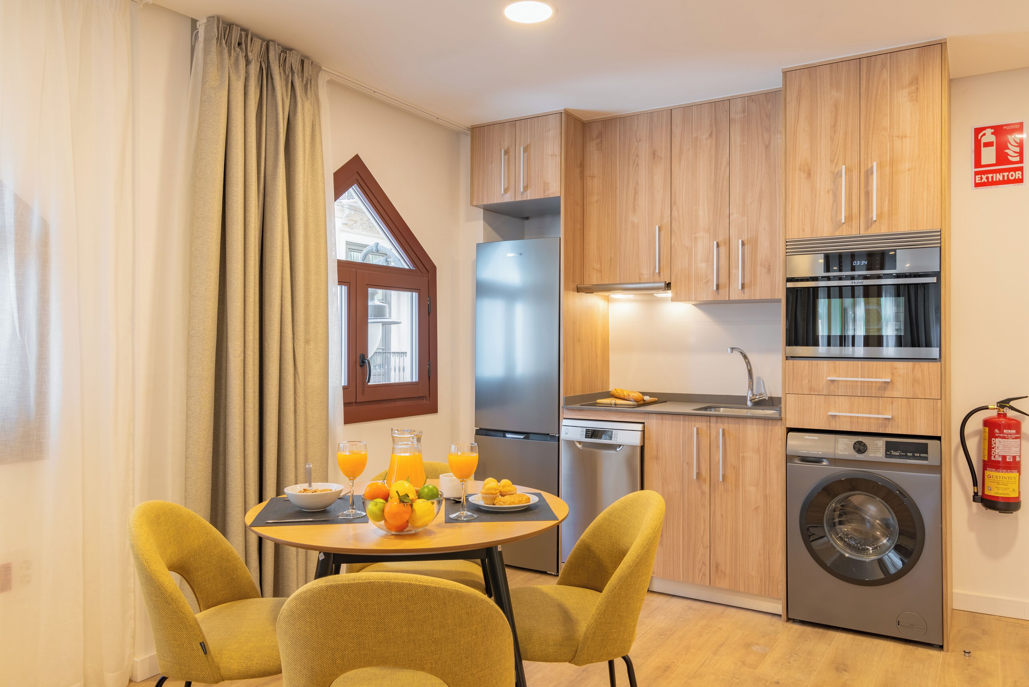 Una cocina moderna con armarios de madera, un refrigerador de acero inoxidable, lavavajillas, lavadora y un comedor con una mesa dispuesta para el desayuno.