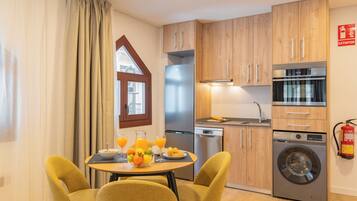 A modern kitchen with wooden cabinets, a stainless steel refrigerator, a dishwasher, a washing machine, and a dining area with a table set for breakfast.