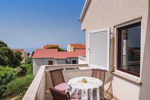 A balcony with a table and chairs, a window with blinds, and a view of red-roofed buildings and greenery.