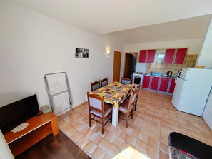 Una cuina compacta amb armaris vermells, una taula de dinar amb cadires, un televisor sobre un suport de fusta i un sòl de rajola.