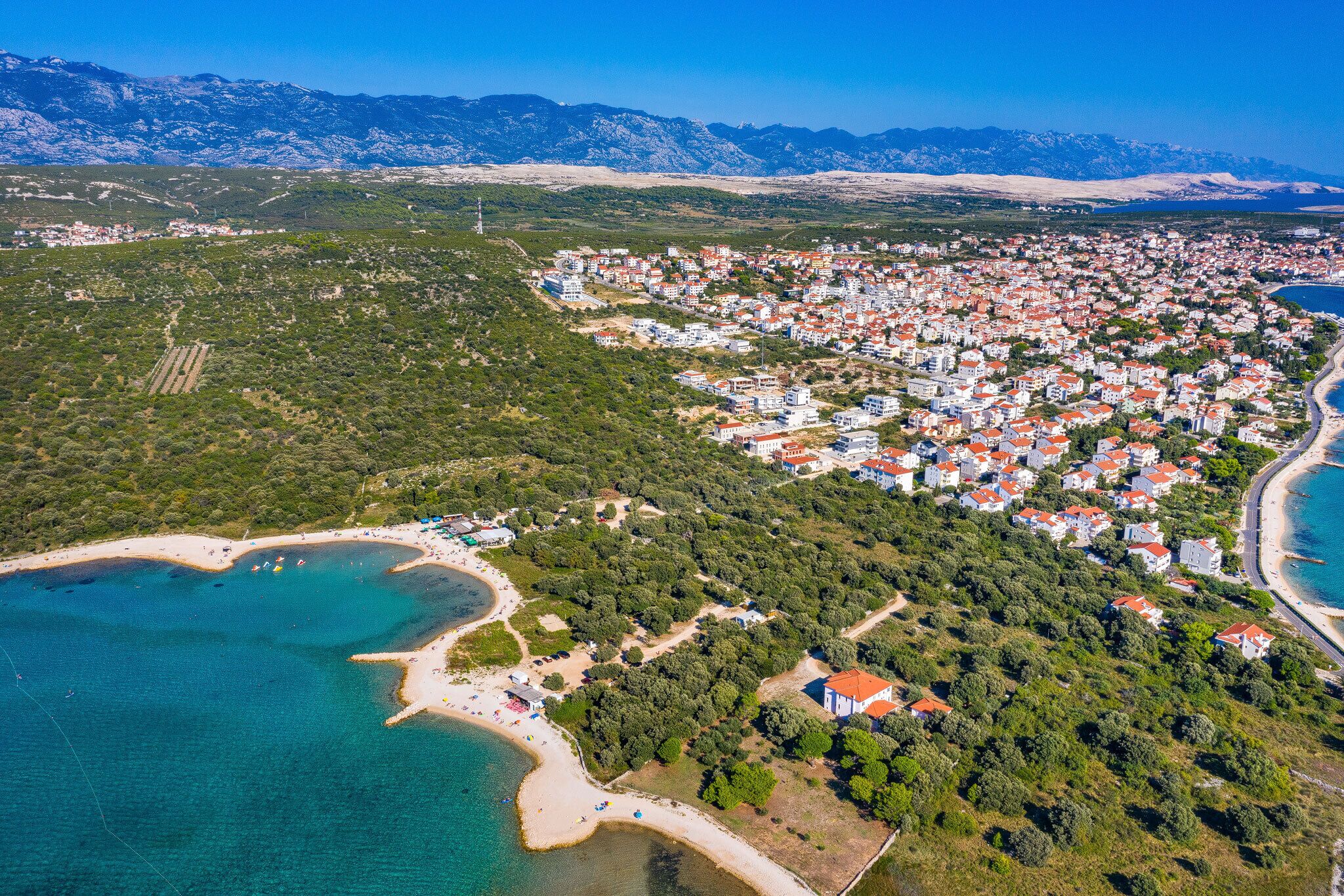 Vue aérienne d’une ville côtière avec une plage, un quartier résidentiel dense et un paysage montagneux en arrière-plan.
