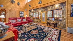 A cozy living room with a red leather sofa, a coffee table, and a large rug. There are stone walls and wooden ceilings, and a stone fireplace.